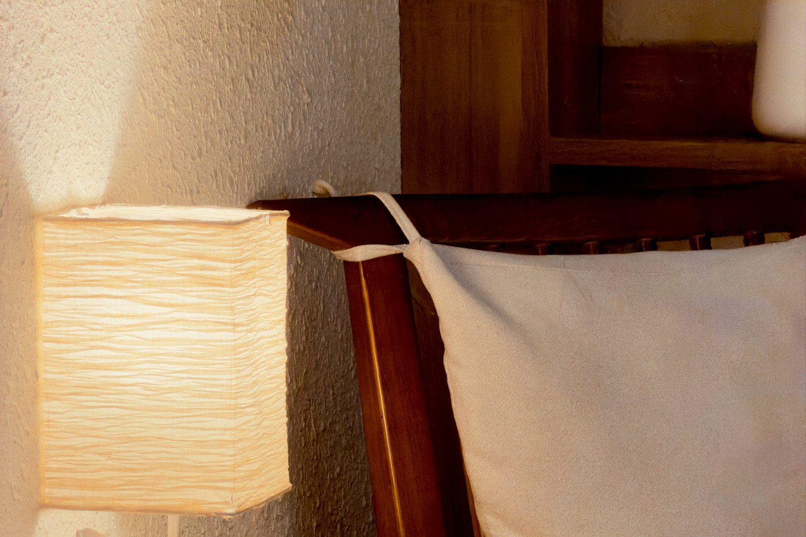 Paper-shaded bedside lamp glowing warm against a textured wall beside a wooden bed frame