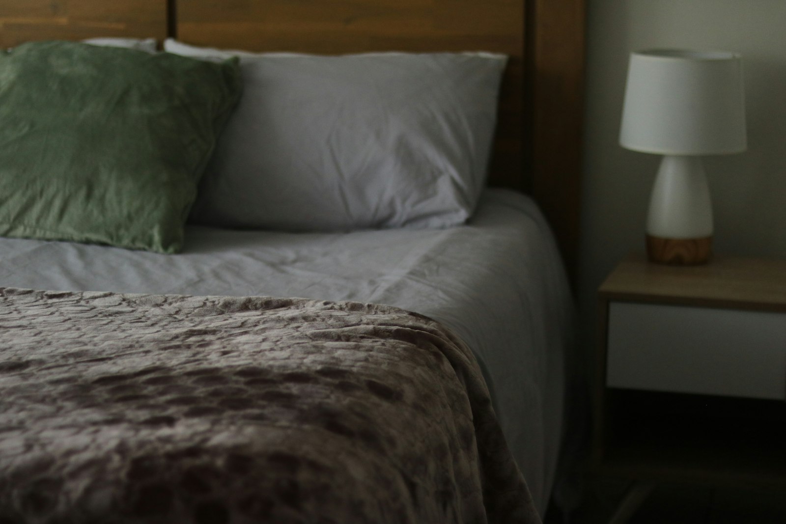 Made bed with gray linens, a textured throw folded at the foot and a small bedside lamp