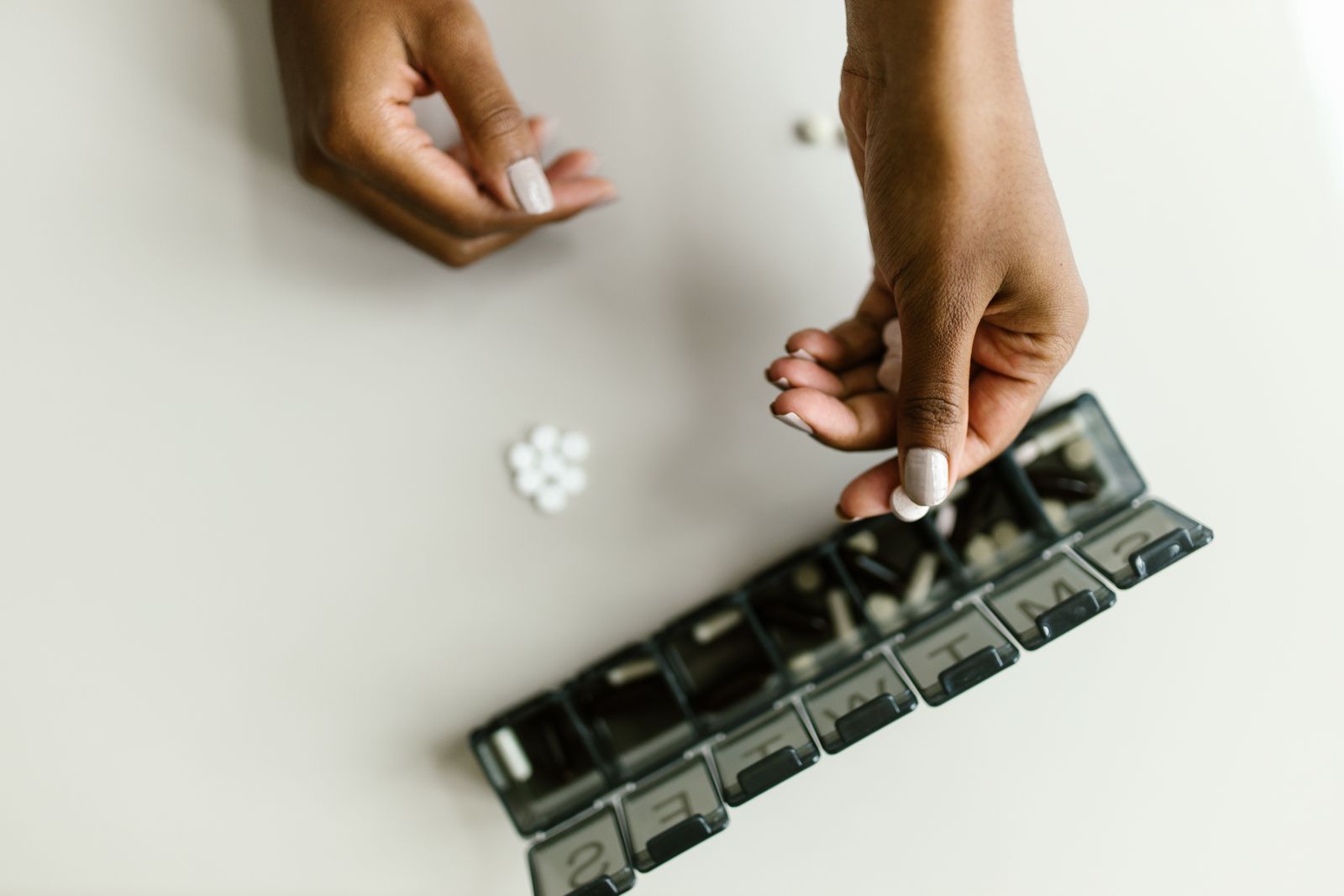 Two hands sorting pills into a weekly compartment organizer on a pale table
