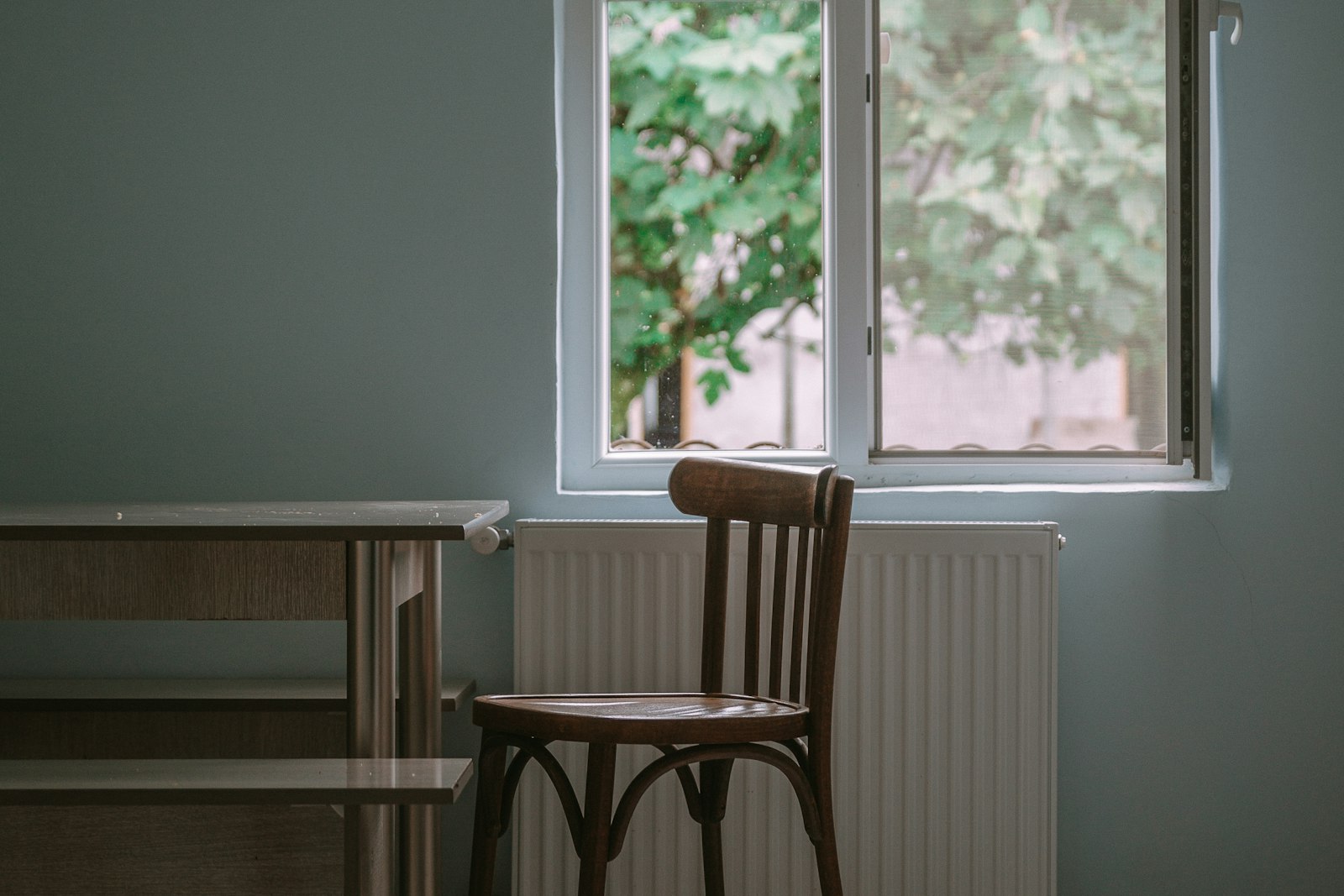 Wooden upright chair with a firm slatted back beside a window and radiator, green leaves outside
