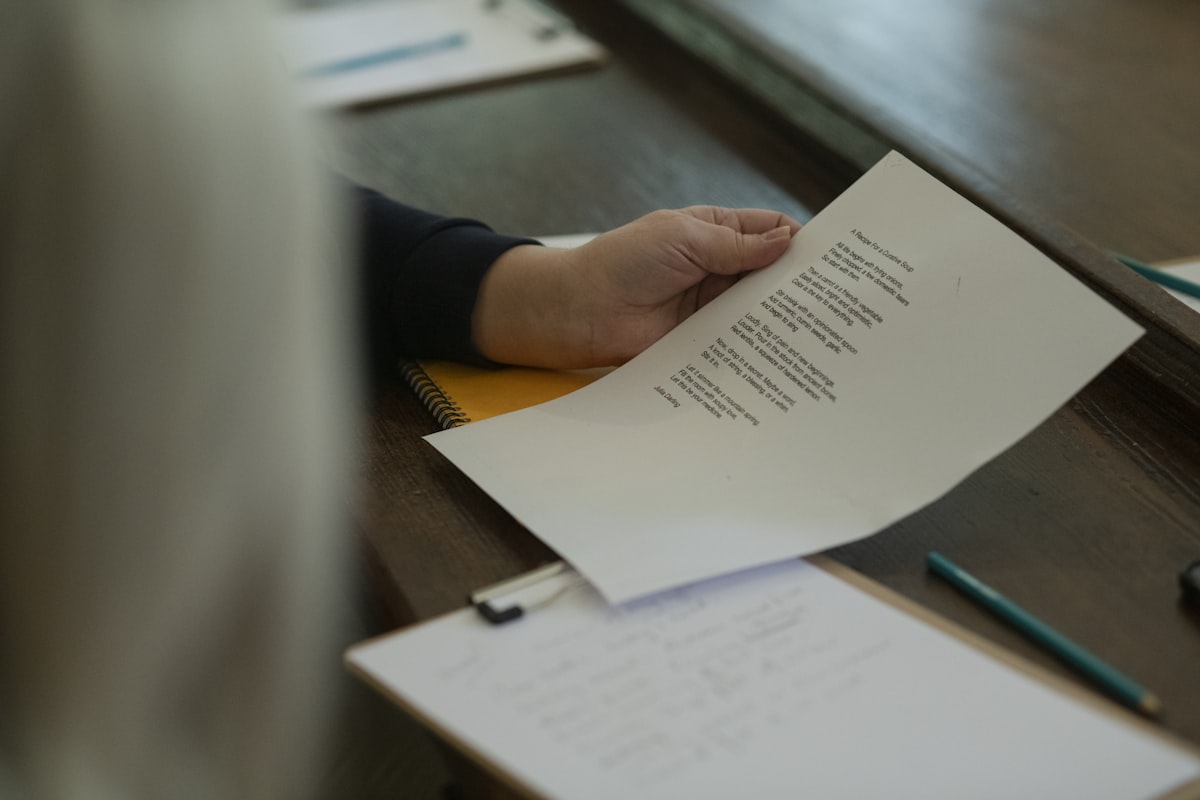 A hand holding a printed document over a wooden table beside a notebook