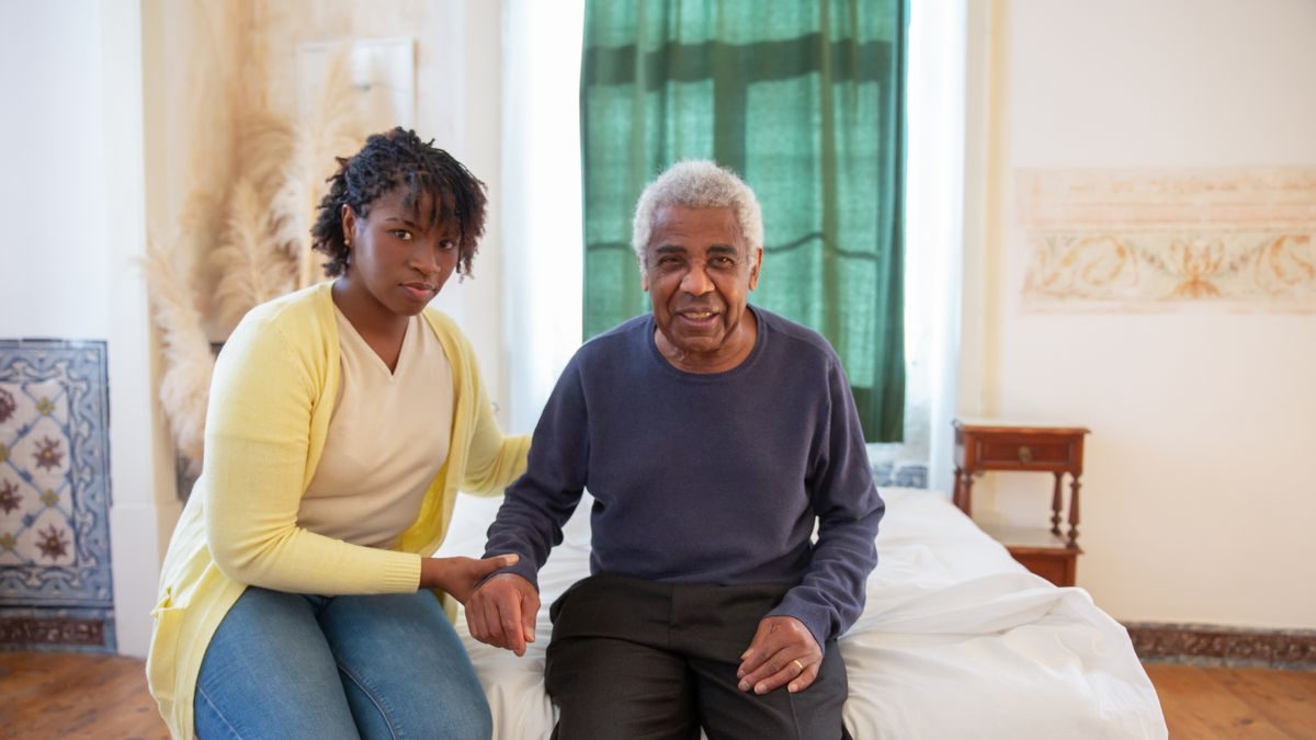 A caregiver sitting with an older man at home, offering a steadying hand