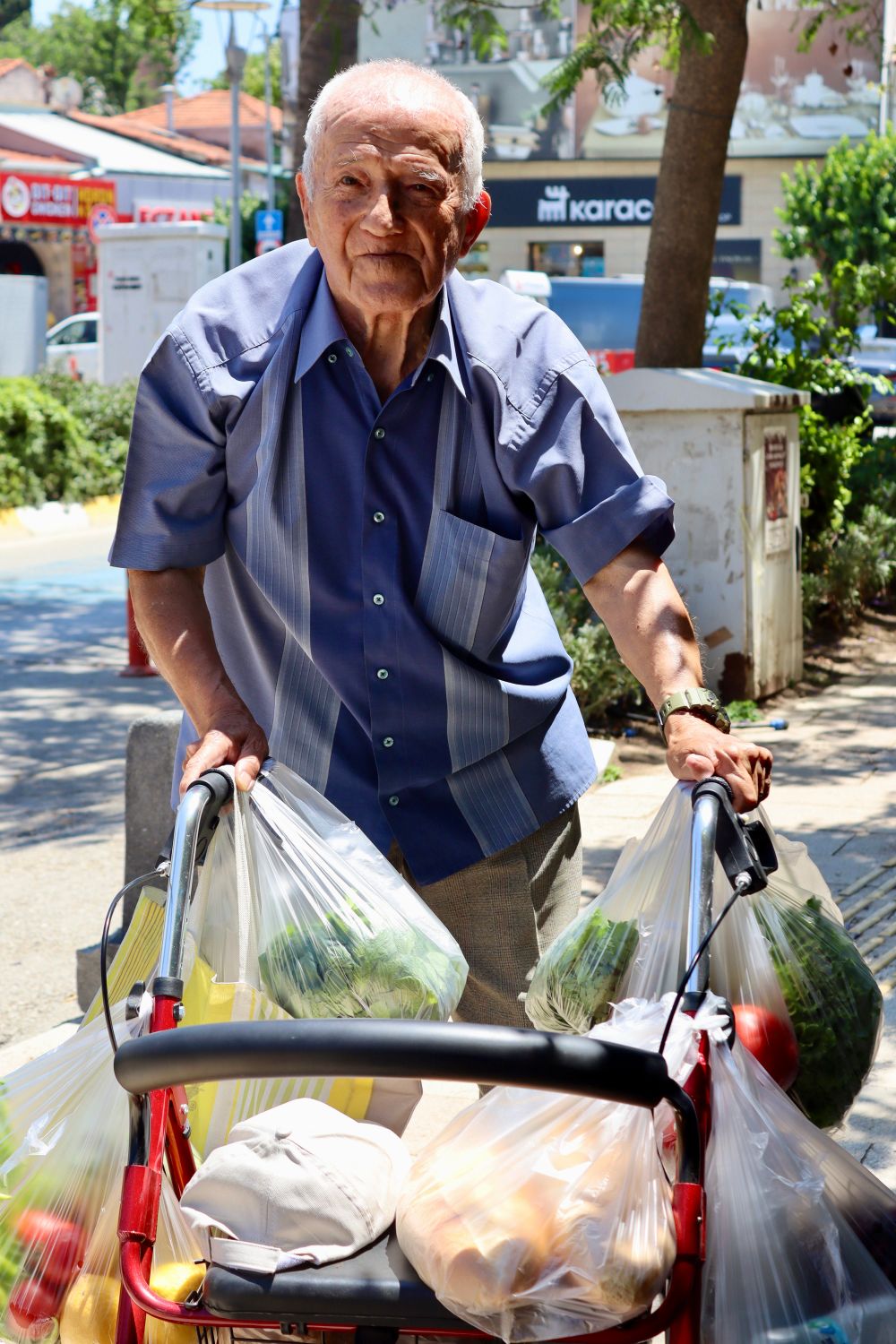 An older man smiling while using a wheeled walker to carry his groceries home on his own